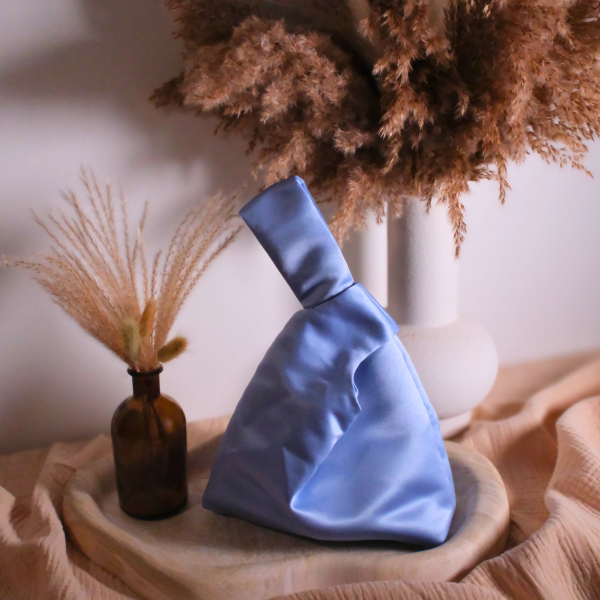 Cornflower blue satin bag knot bag sat on top of a beige marble tray styled with dried grasses and muslin base